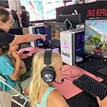 两个小女孩坐在台式电脑前玩《我的世界》(Minecraft)，一个穿着Super Girl Gamer Pro衬衫的女人在旁边帮助她们.